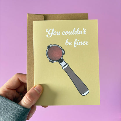 Card with espresso portafilter illustration and text 'You couldn't be finer' held by a hand against a pink background