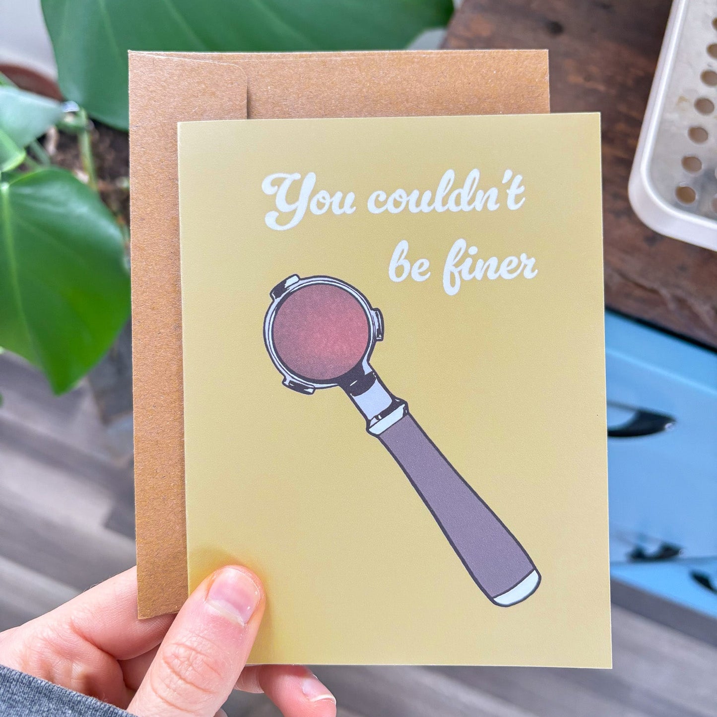 Greeting card with espresso portafilter illustration and text held by a hand, with a plant in the background.