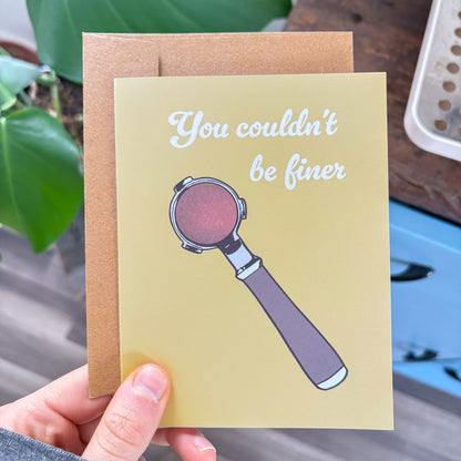 Greeting card with espresso portafilter illustration and text held by a hand, with a plant in the background.