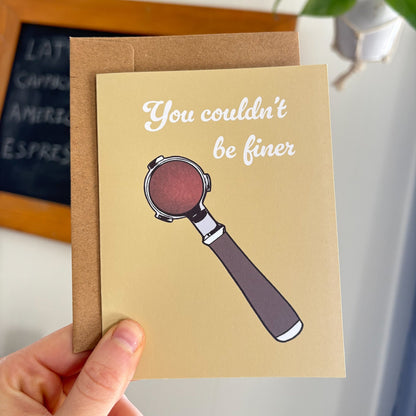 Greeting card with espresso portafilter illustration and text held by a hand, with a plant and chalkboard in the background.