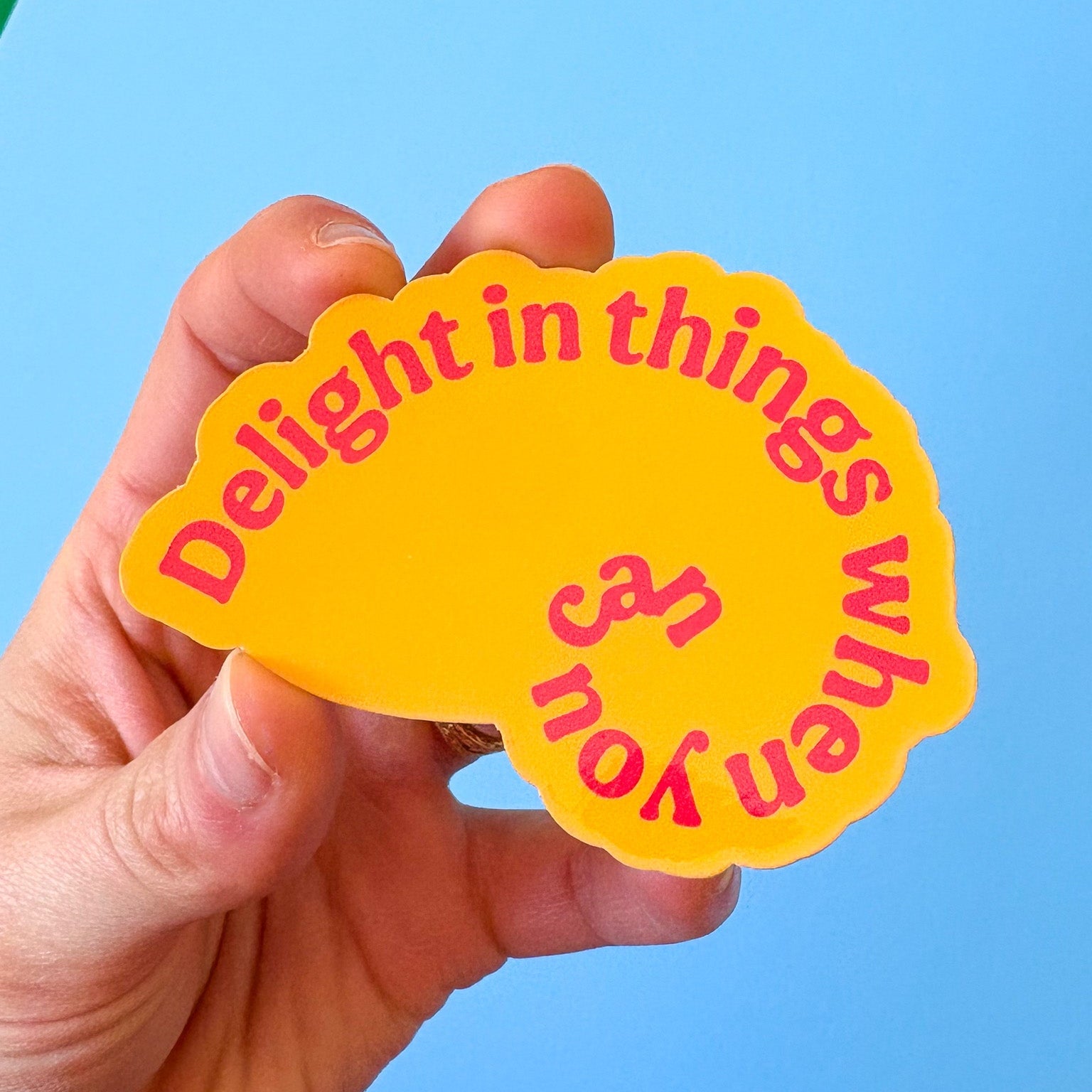Hand holding a shell shaped yellow sticker with red text against a blue background