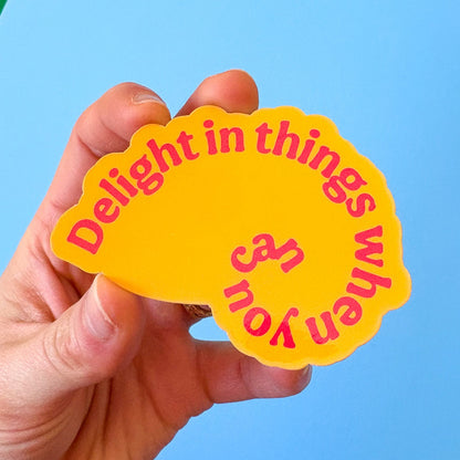 Hand holding a shell shaped yellow sticker with red text against a blue background