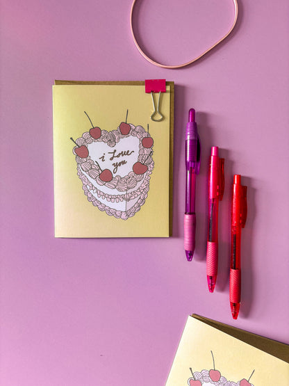Card with a heart-shaped cake illustration and three pens on a pink background.