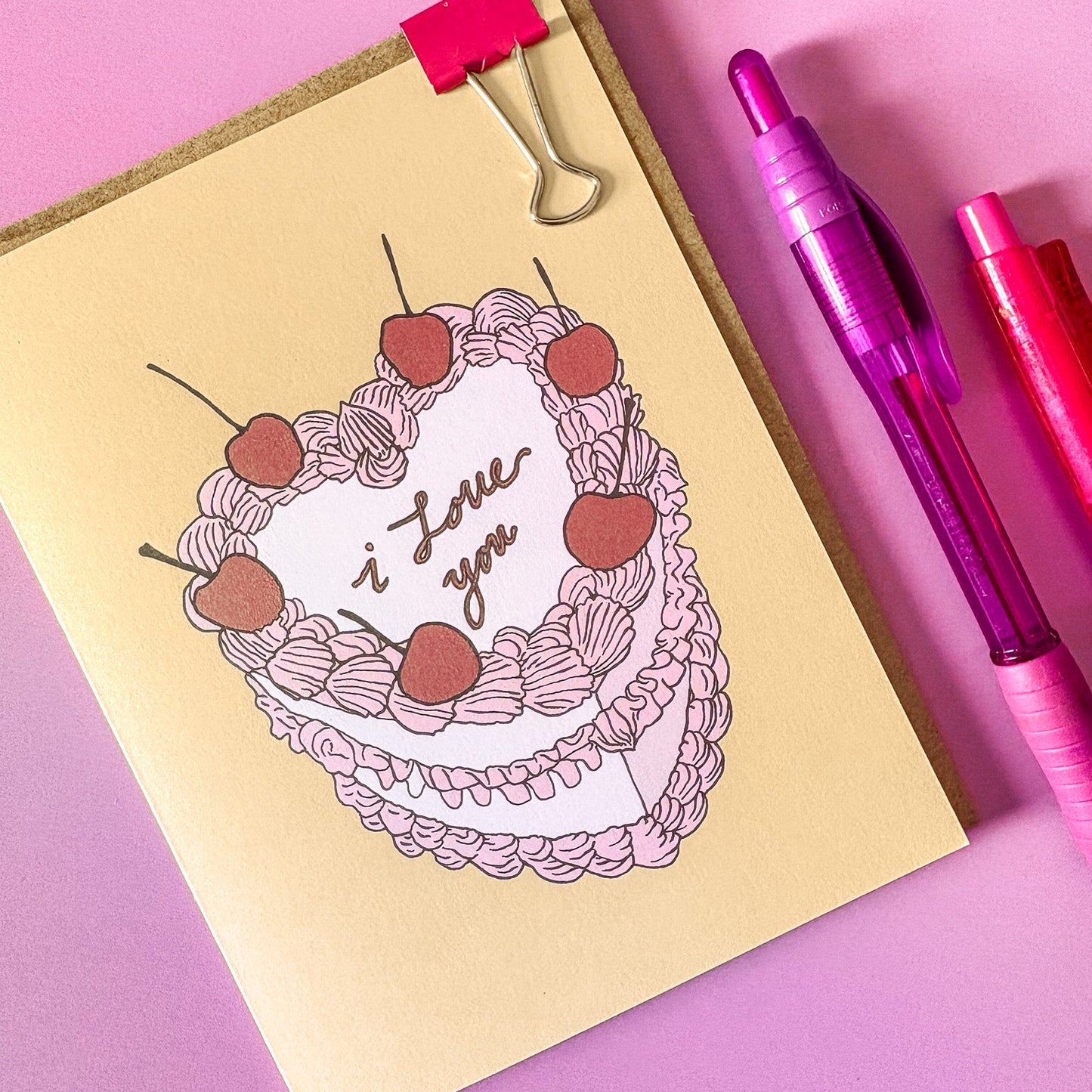 Greeting card with a cake illustration and 'I love you' text on a pink surface with pens.