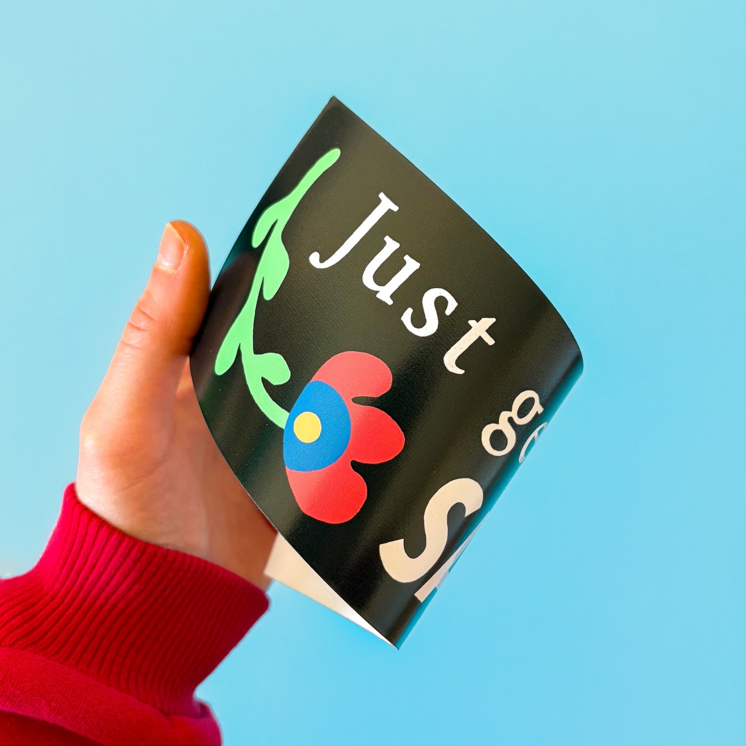 Hand holding a black bumper sticker with a colorful flower design against a blue background.