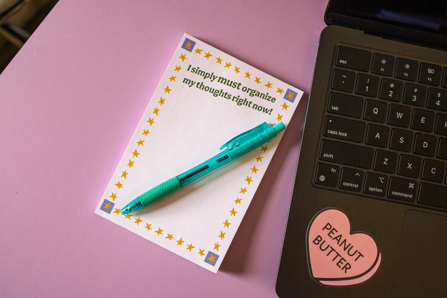 Notepad with a pen on a pink surface next to a laptop.