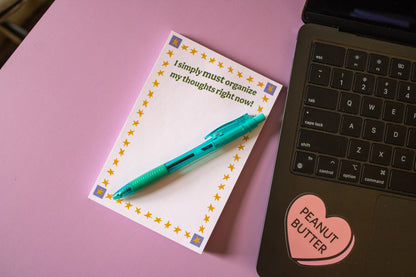 Notepad with a pen on a pink surface next to a laptop.
